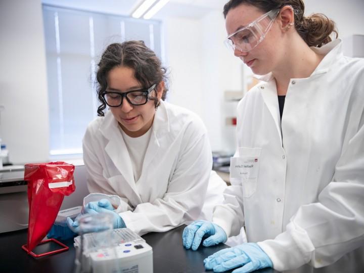 Two students wear goggles, lab coats and gloves while examining their lab project 